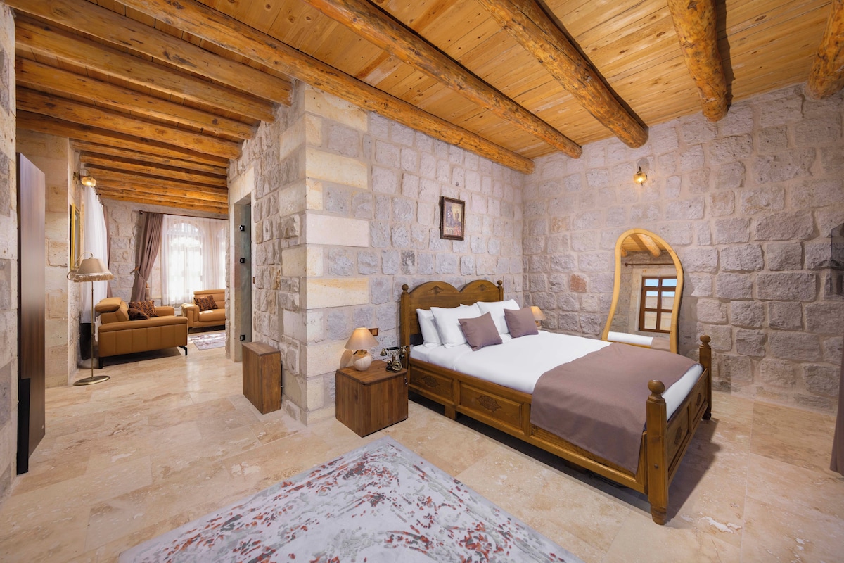 A bedroom with a stone wall, wooden ceiling beams, a bed with a wooden headboard, a nightstand, a mirror, and a window with curtains.