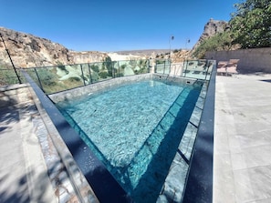 Una piscina rectangular al aire libre con agua cristalina, rodeada por una barandilla de vidrio, con un paisaje rocoso de fondo.
