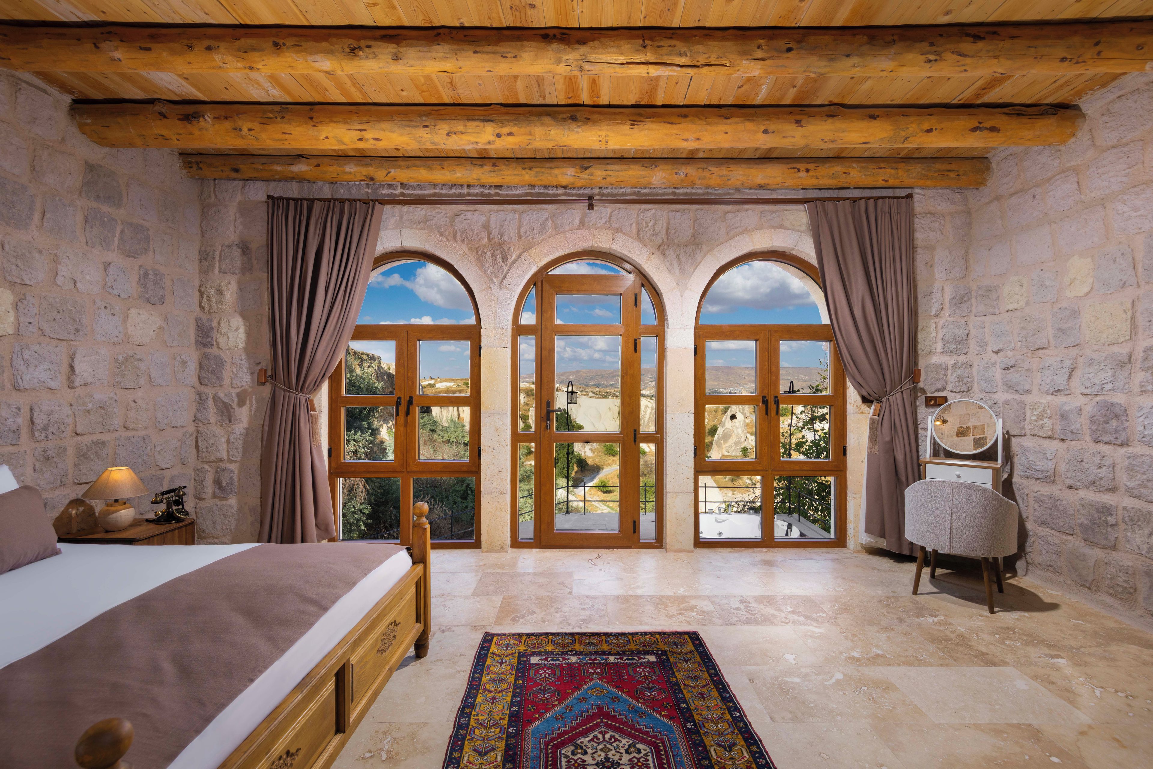 A bedroom with a stone wall, a wooden ceiling, a bed, a nightstand, a lamp, a chair, and a view through arched windows.