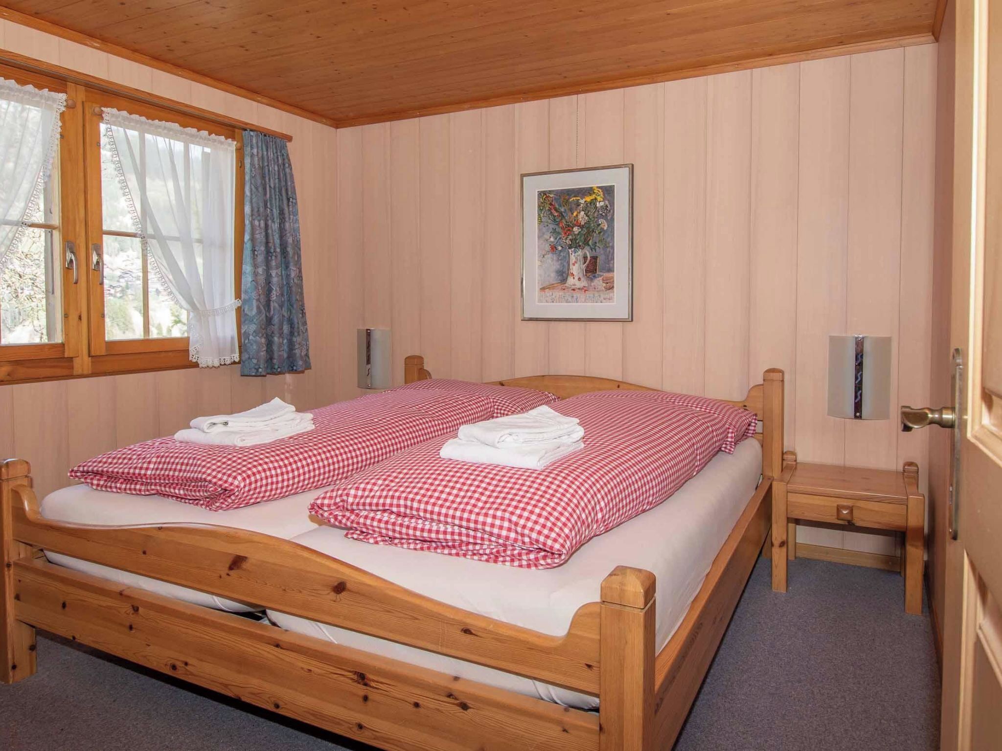 A wooden bed with checkered bedding, a bedside table, a nightstand, a window with curtains, and a framed picture on the wall.