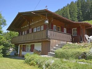 A two-story wooden chalet with a balcony, surrounded by greenery and a mountainous backdrop. - Chalet Iris Appartement/Fewo, Dusche, WC, 2 Schlafräume 3-Zimmerwohnung by Interhome (Adelboden)
