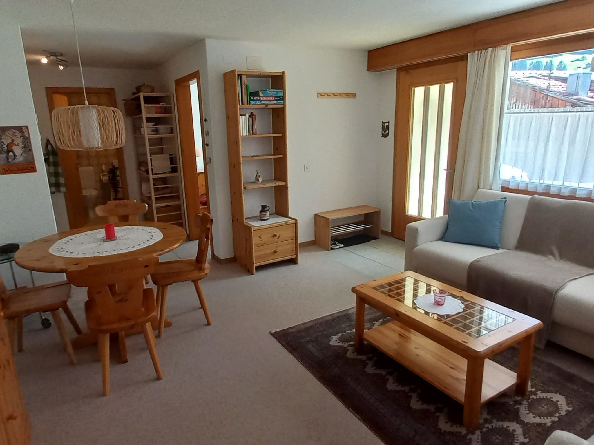 A living room with a sofa, a coffee table, a dining table with chairs, a bookshelf, and a window with curtains.