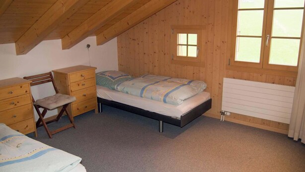 Una camera da letto con soffitto a falde, pareti in legno, un letto con copripiumino a motivi, una scrivania con cassetti, una sedia e una finestra con vista.