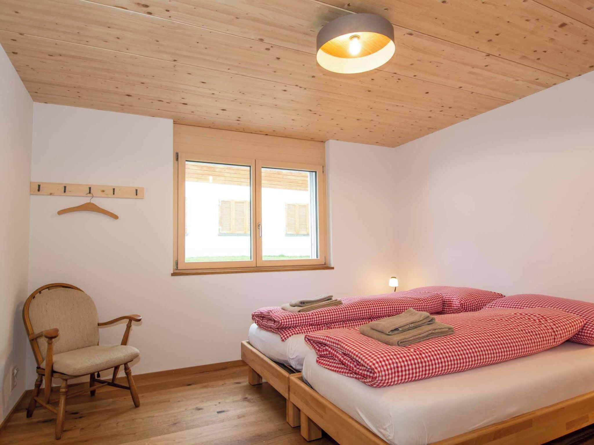 Ein Zimmer mit Holzboden, einem Bett mit rot-weiß karierter Bettwäsche, einem Sessel und einem Fenster mit Rollläden.
