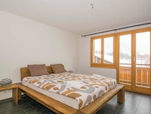 A bedroom with a wooden bed, a nightstand, a large window with a view of a snowy landscape, and a ceiling light. - Schützenrain 1. Stock Appartement/Fewo, Bad, WC, 3 Schlafräume by Interhome (Adelboden)