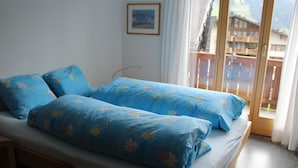 A bed with floral bedding, a window with a view of buildings, and a picture on the wall. - Haus Baumann Appartement/Fewo, Bad, WC, 4 oder mehr Schlafräume 5 Zimmerwohnung by Interhome (Adelboden)