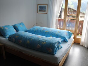 A bed with floral bedding, a window with a view of buildings, and a picture on the wall. - Haus Baumann Appartement/Fewo, Bad, WC, 4 oder mehr Schlafräume 5 Zimmerwohnung by Interhome (Adelboden)