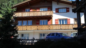 A multi-story building with a wooden balcony, a car parked in front, and a mountainous backdrop. - Haus Baumann Appartement/Fewo, Bad, WC, 4 oder mehr Schlafräume 5 Zimmerwohnung by Interhome (Adelboden)