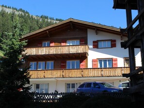 A multi-story building with a wooden balcony, a car parked in front, and a mountainous backdrop. - Haus Baumann Appartement/Fewo, Bad, WC, 4 oder mehr Schlafräume 5 Zimmerwohnung by Interhome (Adelboden)