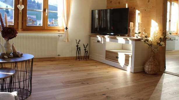 A modern living room with a large flat-screen TV, wooden flooring, a dining table with chairs, and a view of the outdoors through the windows. - Flott Appartement/Fewo, Dusche und Bad, WC, 1 Schlafraum 2.5-Zimmerwohnung by Interhome (Adelboden)