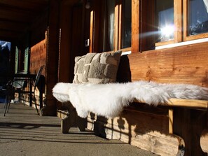 Un banc en bois recouvert d’un plaid en fourrure, un mur de pierre et une fenêtre baignée de lumière.