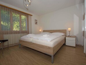 A neatly made bed with white linens, a bedside table with a lamp, and a window with patterned curtains. - Helios Appartement/Fewo, Dusche, WC, 3 Schlafräume 4-Zimmerwohnung by Interhome (Adelboden)