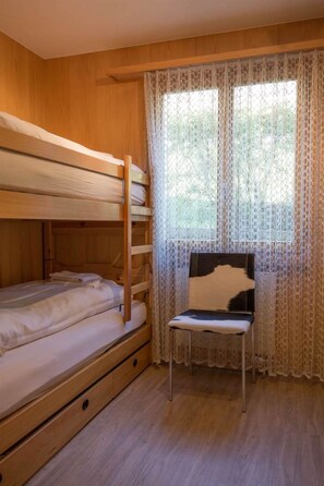 A bunk bed room with a wooden floor, a chair with a black and white headrest, and lace curtains on the window. - Helios Appartement/Fewo, Dusche, WC, 3 Schlafräume 4-Zimmerwohnung by Interhome (Adelboden)