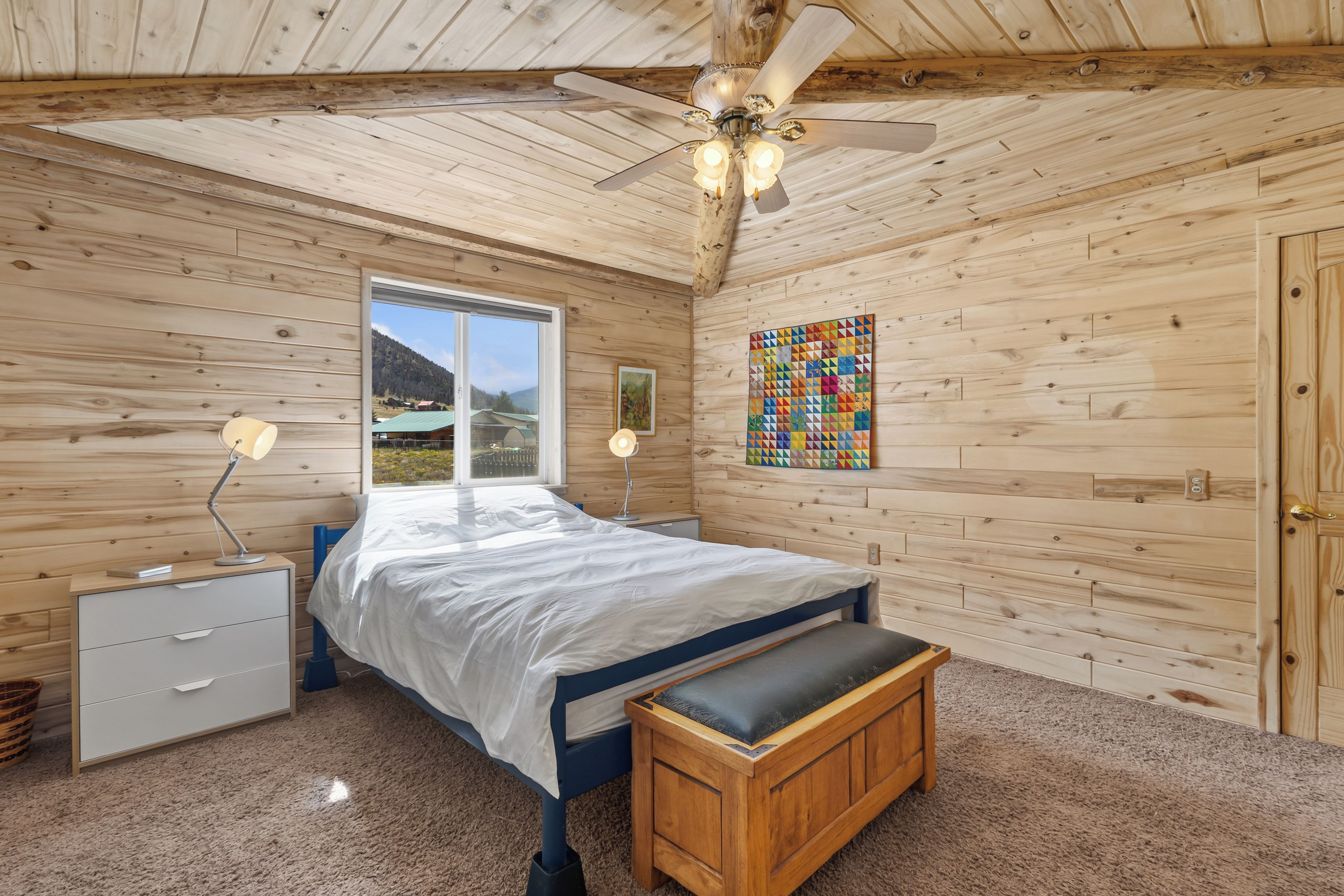 Un dormitorio con cama, mesitas de noche, un banco, ventilador de techo y ventana con vistas, además de una obra de arte colorida en la pared.