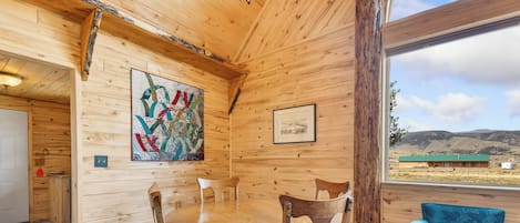 A wooden interior with a round table and chairs, a framed picture, and a view of the outdoors.