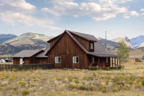 A rustic wooden building with a sloped roof, surrounded by a fenced yard and a mountainous backdrop.
