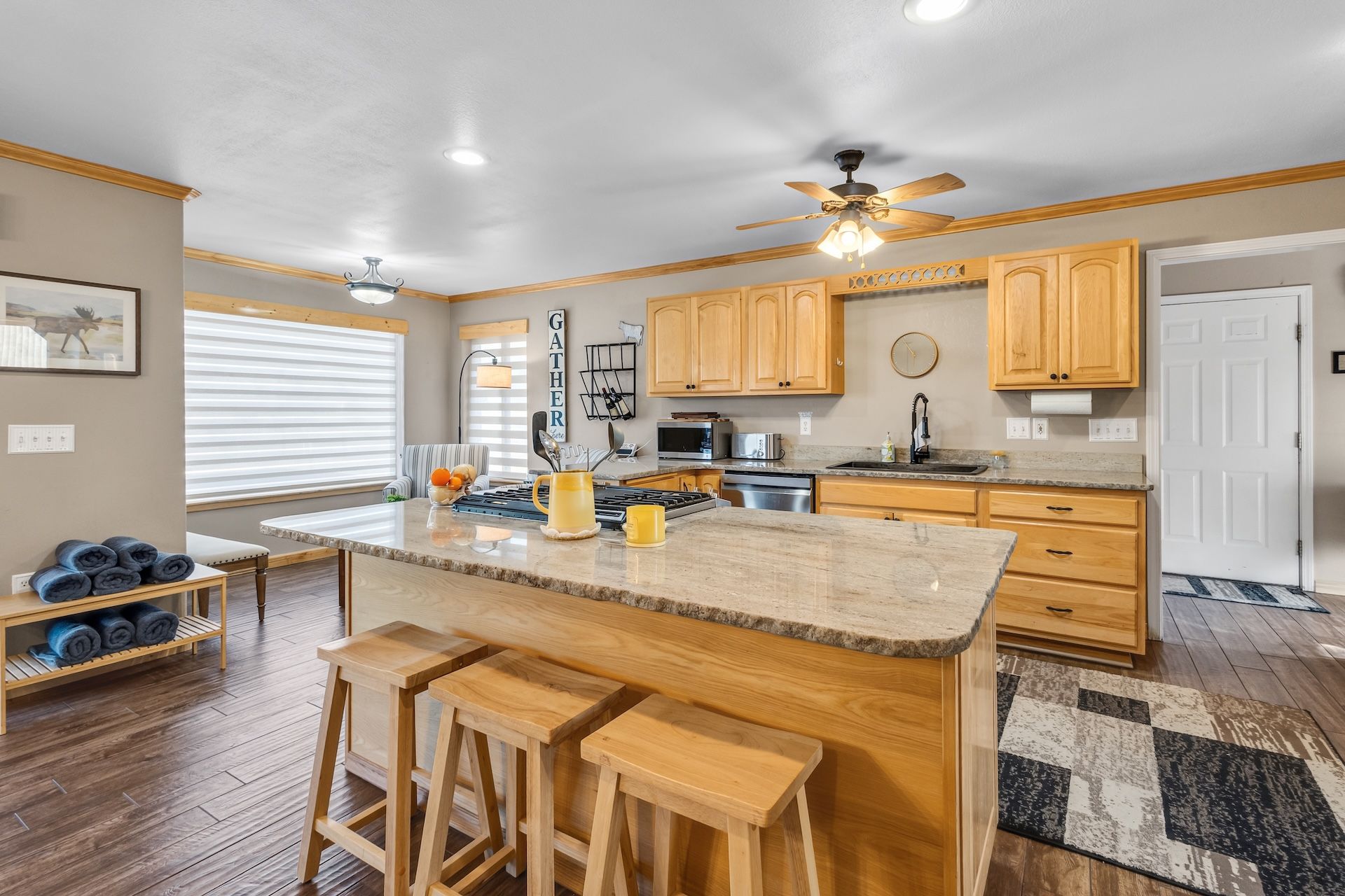 A modern kitchen with wooden cabinets, a central island with stools, a ceiling fan, and a view of the living area.