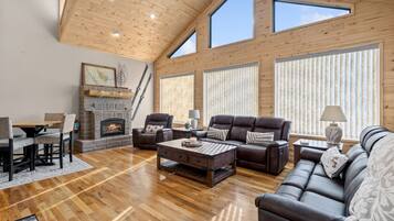 A spacious living room with a fireplace, leather sofas, wooden flooring, and large windows.