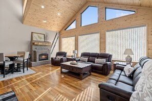 A spacious living room with a fireplace, leather sofas, wooden flooring, and large windows.