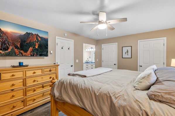 Quarto com cama, cômoda, ventilador de teto e pintura de paisagem de montanha.
