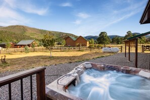 Een hottub met een houten terras in een landelijke omgeving, met huizen en bergen op de achtergrond.