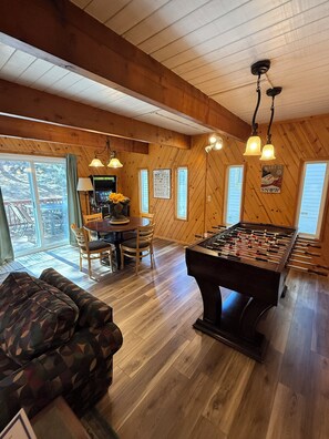 A cozy room with a foosball table, wooden walls, and a dining area with a table and chairs. - Unique Big Bear Experience, Family Game Room, plenty of space to lounge - BEST location to everything! Hot tub and huge yard, great for sledding. So close to downtown - walk to The Village for boutique shopping! (Big Bear Lake)