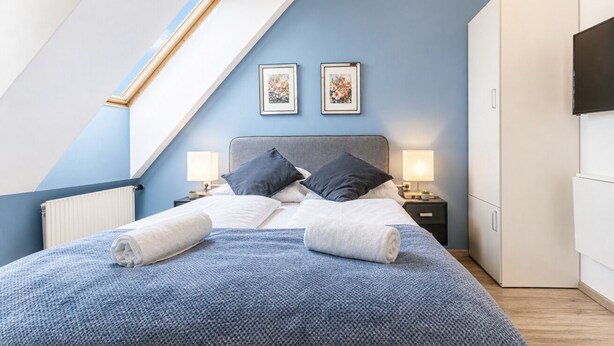 A bedroom with a sloped ceiling, a bed with two pillows, a nightstand with a lamp, and a wardrobe.