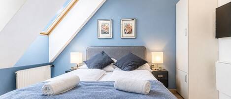 A bedroom with a sloped ceiling, a bed with two pillows, a nightstand with a lamp, and a wardrobe.