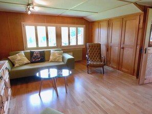 A room with a wooden floor, a green sofa with cushions, a round glass table, a brown armchair, and a large window with multiple panes. - Stockbrunä Appartement/Fewo, Bad, WC, 2 Schlafräume by Interhome (Adelboden)