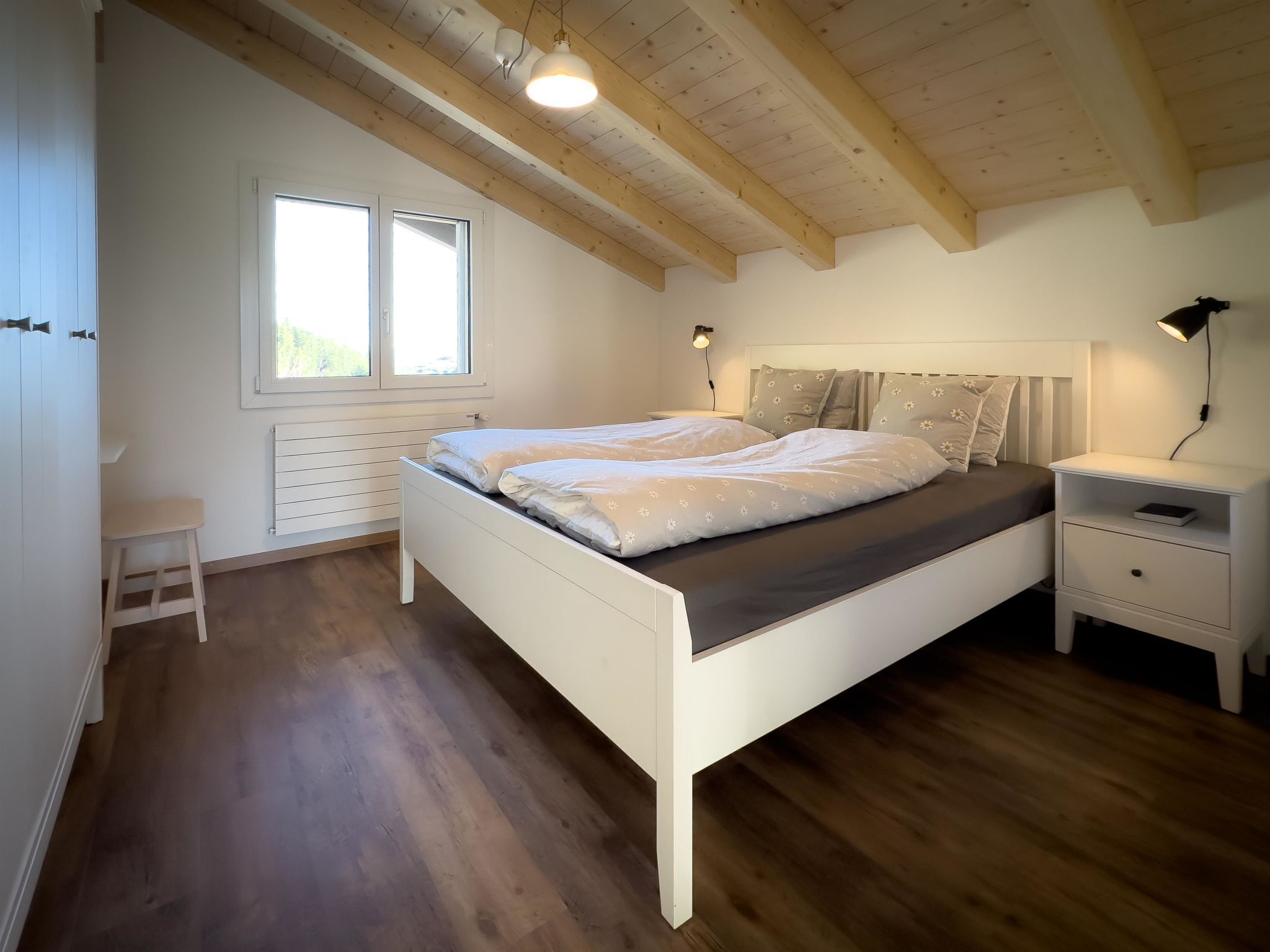 Une chambre avec un lit blanc, une table de chevet, un petit tabouret, une fenêtre et des poutres en bois au plafond.