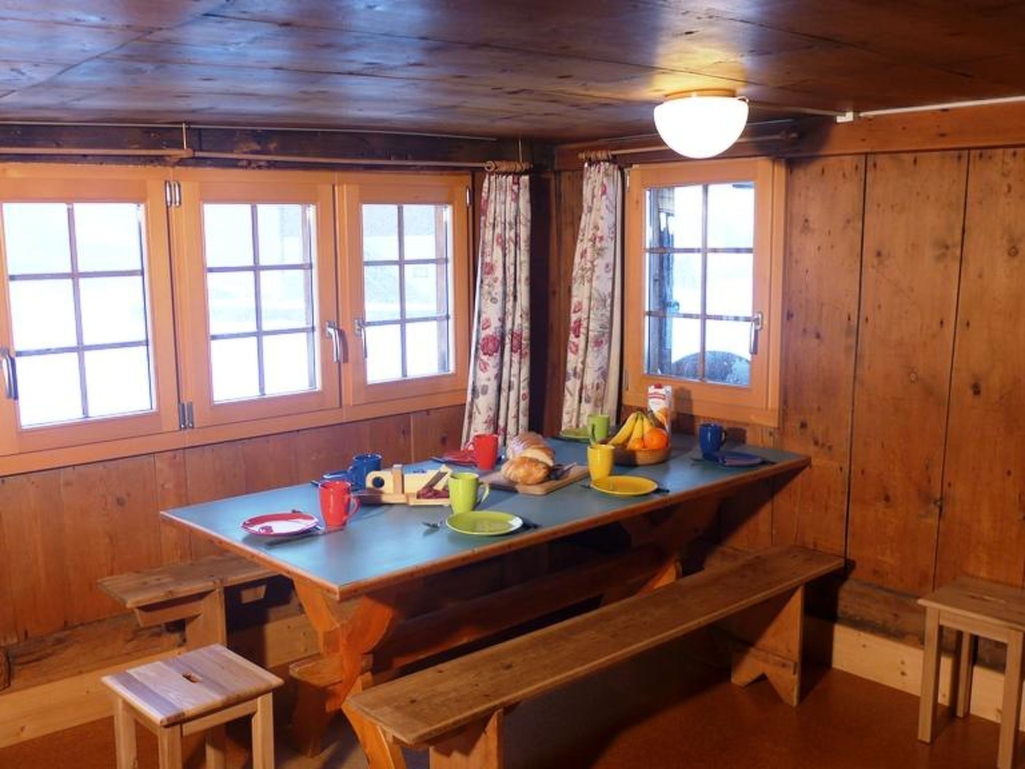 A wooden dining area with a large table, benches, and windows with curtains.