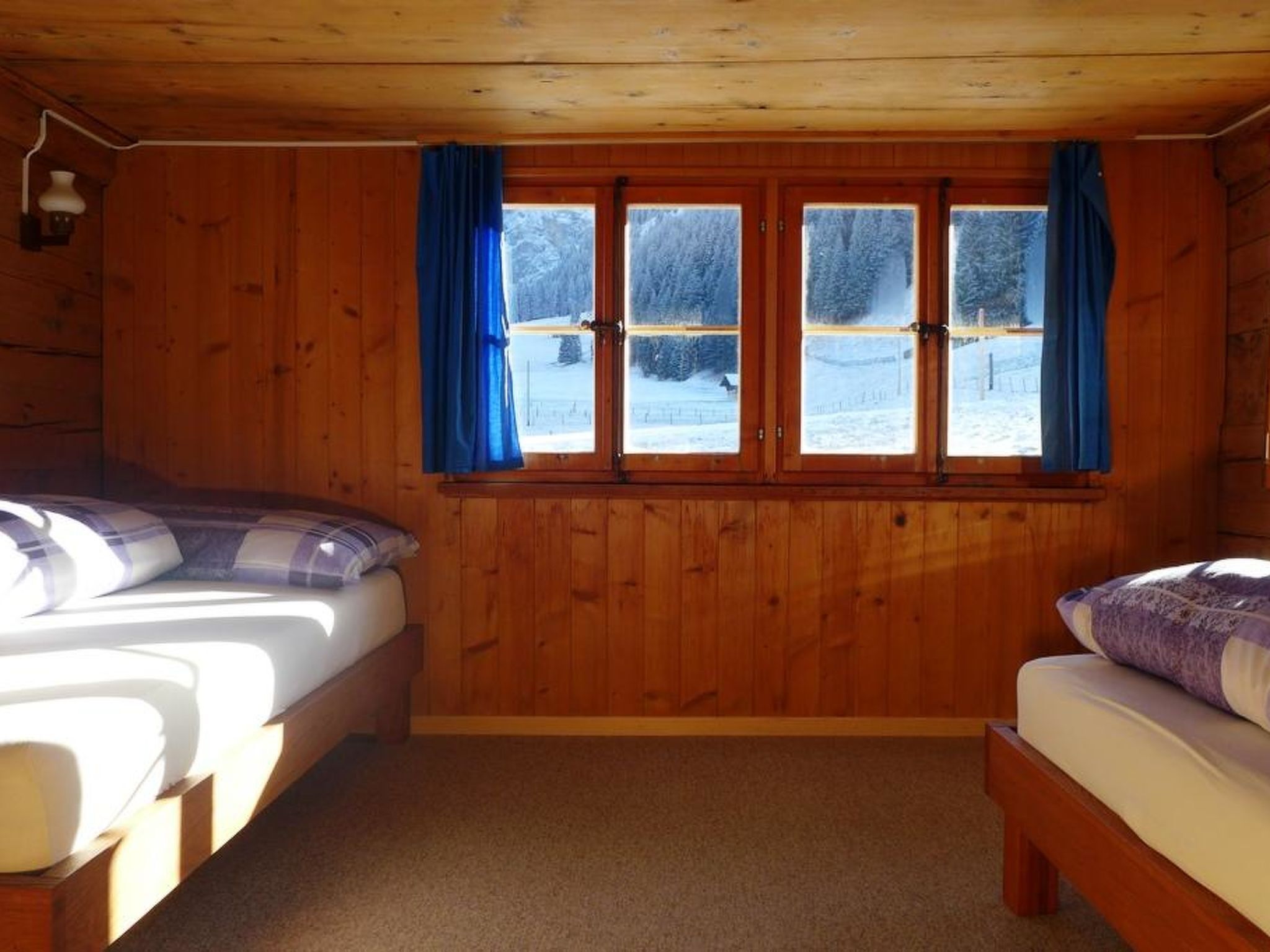 A cabin with two beds, wooden walls, a window with curtains, and a view of a snowy landscape.