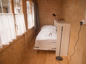 Ein kleines Zimmer mit einem Einzelbett, Holzwänden und einem Fenster mit Vorhängen.