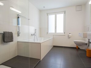 Ein modernes Badezimmer mit einer Glaskabinentrennung zur Badewanne, einer Toilette und einem Fenster.