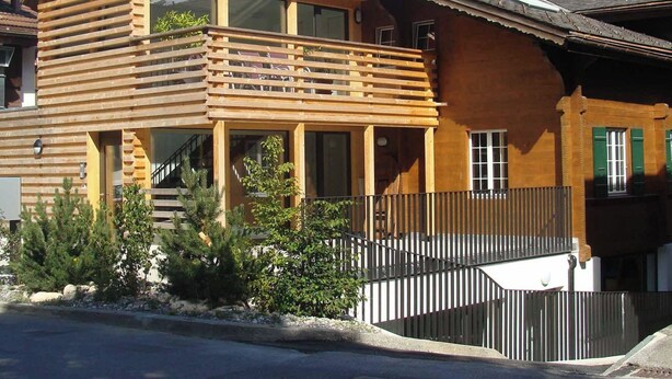 Eine Holzhütte mit Balkon, großen Fenstern und einem Bergpanorama im Hintergrund.