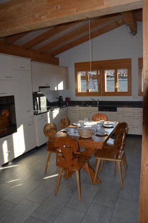 Ein Essbereich mit einem Holztisch, gedeckt für vier Personen, umgeben von Stühlen. Im Hintergrund ist eine Küchenzeile mit Schränken und einem Herd zu sehen.