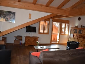 Ein gemütliches Wohnzimmer mit schrägem Dach, Holzmöbeln, einem Fernseher und Blick ins Freie.