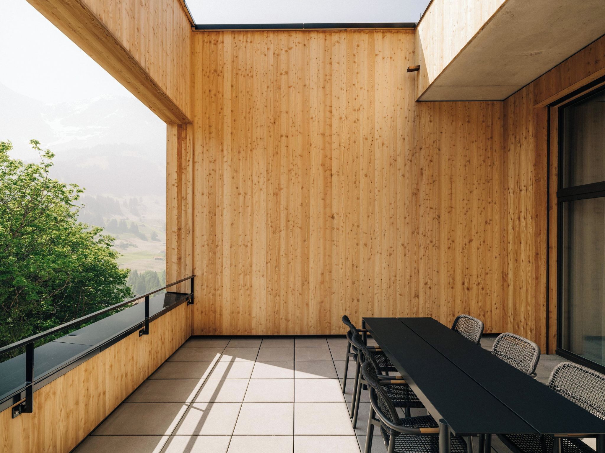 Un balcón de madera con una mesa y sillas, barandilla de vidrio y vistas a la vegetación.