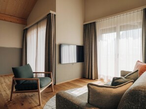 Ein Wohnzimmer mit Holzboden, einem an der Wand befestigten Flachbildfernseher, einem grünen Sessel, einer Couch und einem gemusterten Teppich.