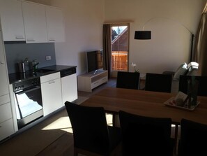 Eine moderne Küche mit weißen Schränken, einer schwarzen Arbeitsplatte und einem eingebauten Ofen. Ein Essbereich mit einem Holztisch und schwarzen Stühlen ist ebenfalls vorhanden. Ein Fernseher ist an der Wand montiert, und eine Lampe steht auf dem Tisch.