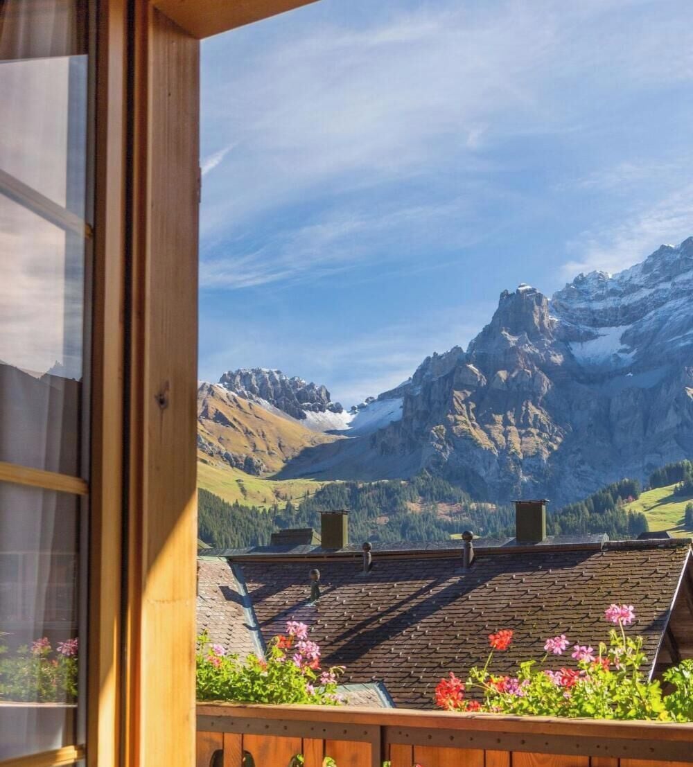 Blick aus einem Fenster auf eine gebirgige Landschaft mit einem Holzbalkon und Blumentöpfen.