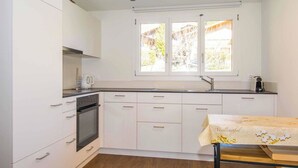 Eine kleine Küche mit weißen Schränken, einem eingebauten Ofen, einem Fenster mit Blick ins Grüne und einem Tisch mit Tischdecke.