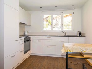A compact kitchen with white cabinets, a built-in oven, a window with a view of greenery, and a small table with a tablecloth. - Chalet Buchhüttli Appartement/Fewo, Dusche oder Bad, WC, 2 Schlafräume by Interhome (Adelboden)