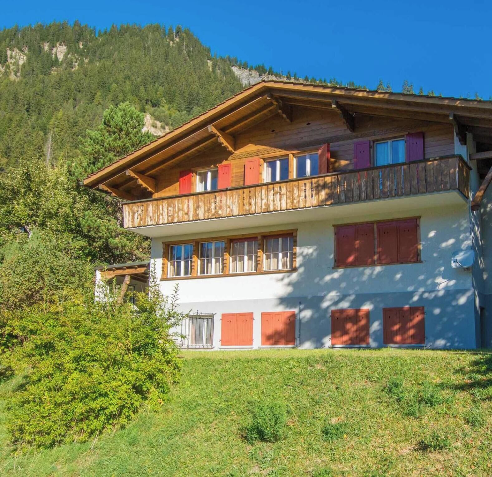 A two-story chalet with a balcony, set against a mountainous backdrop.