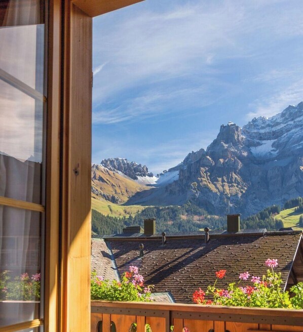 Vista desde una ventana con vistas a un paisaje montañoso, con un balcón de madera y macetas con flores.