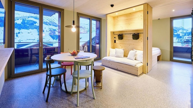 A modern interior with a dining area, a sofa, and a view of snowy mountains through large windows. - Apart Hotel Adelboden am Dorfplatz Appartement/Fewo, Dusche, WC by Interhome (Adelboden)