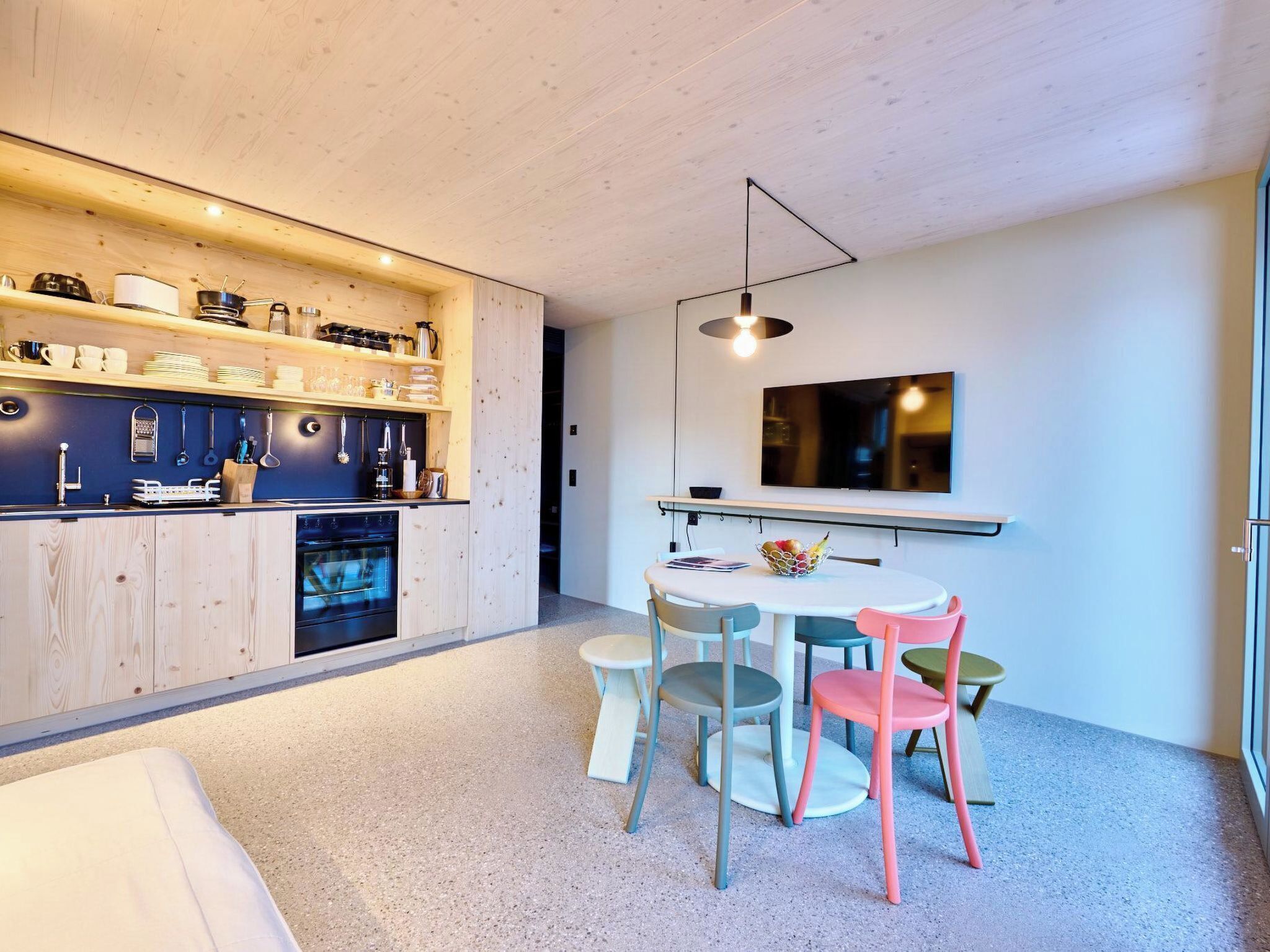 A modern kitchen with wooden cabinets, a built-in oven, and a dining area with a round table and colorful chairs.
