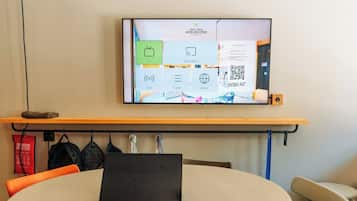 Ein Laptop steht auf einem runden Tisch, neben ihm liegen eine Fernbedienung und ein Notizblock. An der Wand montiert ist ein Fernseher, der einen QR-Code und verschiedene Symbole zeigt.