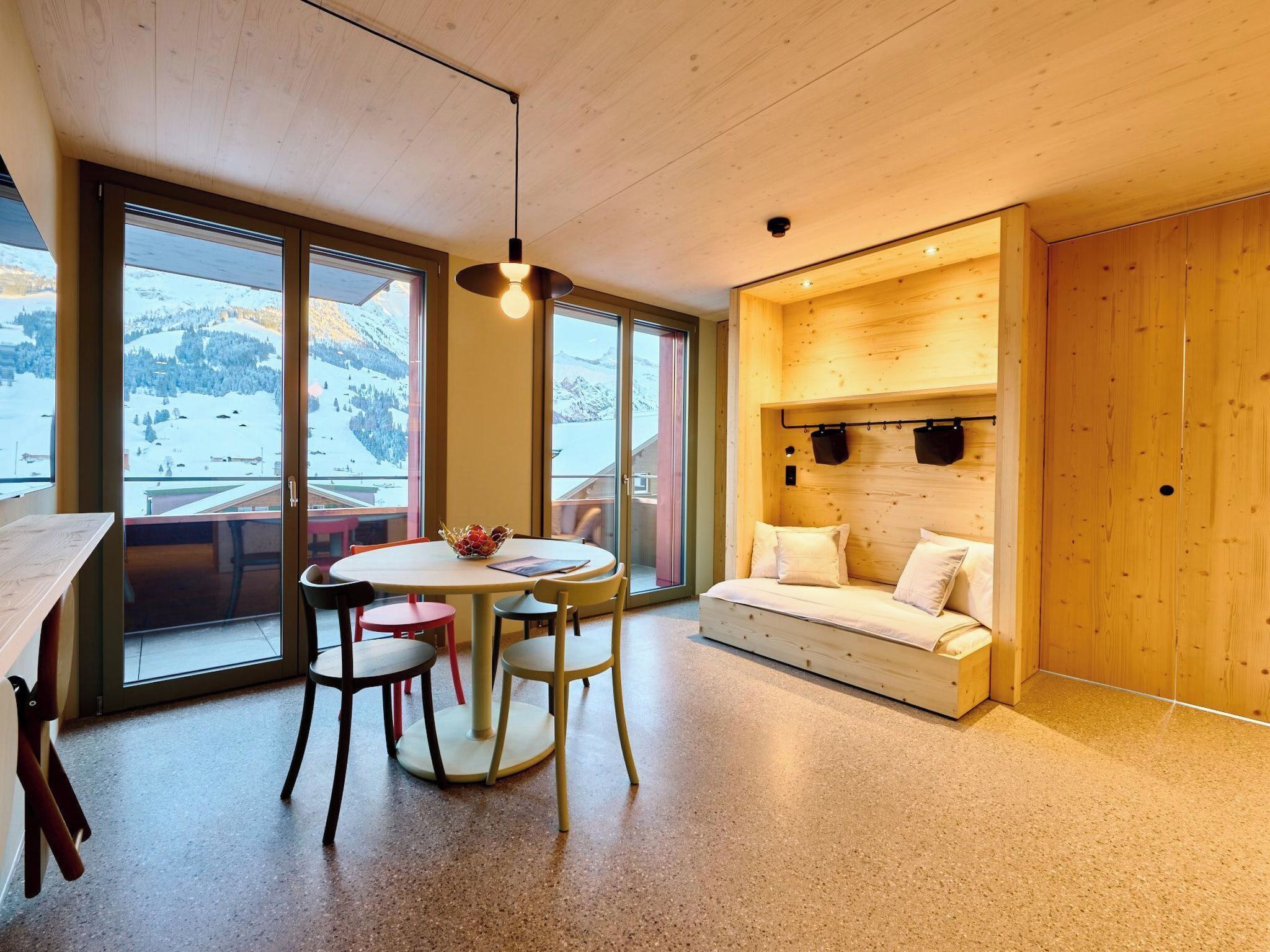 Un intérieur moderne avec un plafond en bois, un coin repas comprenant une table ronde et des chaises, un canapé avec des coussins et de grandes fenêtres offrant une vue sur un paysage enneigé.