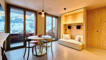 Un intérieur moderne avec un plafond en bois, un coin repas comprenant une table ronde et des chaises, un canapé avec des coussins et de grandes fenêtres offrant une vue sur un paysage enneigé.